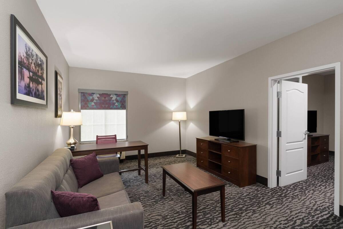 A hotel room with a sofa, a table, a TV, a lamp, and a window with a geometric pattern.