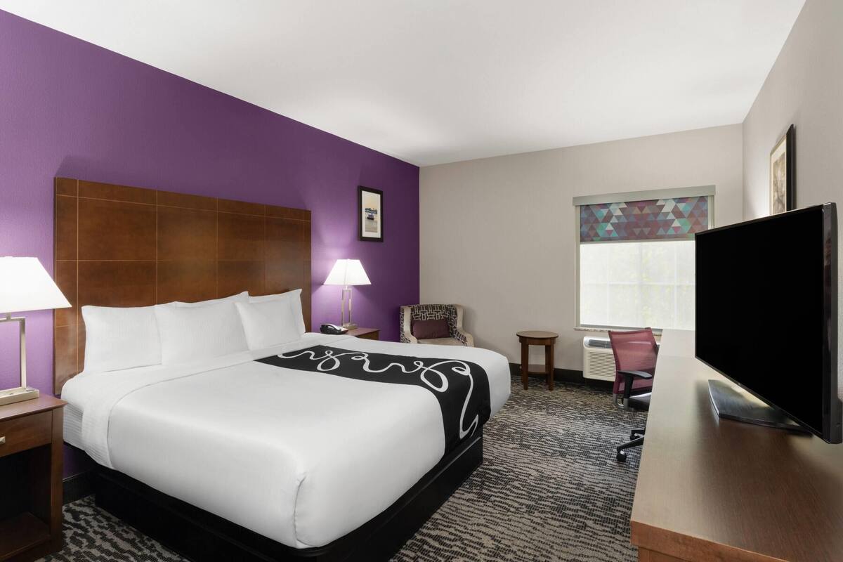 A hotel room with a large bed, a desk with a flat-screen TV, a chair, a lamp, and a window with blinds.