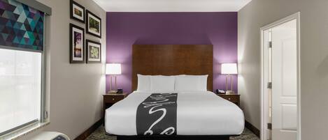 A hotel room with a bed, a window, a wall-mounted air conditioner, a nightstand, a lamp, and a door.