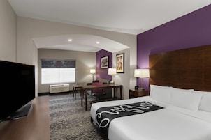 Habitación de hotel con cama, escritorio, silla, televisor y ventana con persianas.