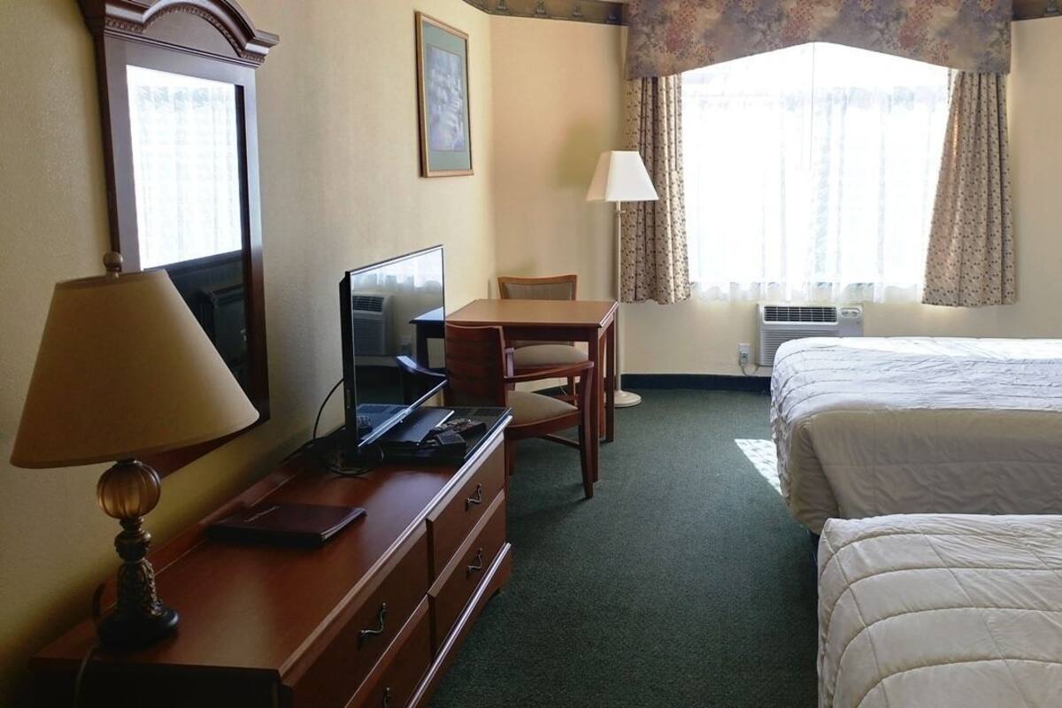 Ein Hotelzimmer mit Bett, Schreibtisch mit Computer, Stuhl, Lampe und einem Fenster mit Vorhängen.