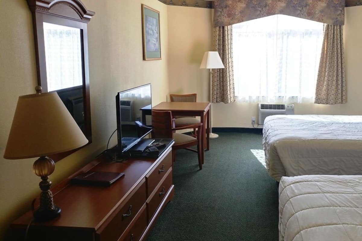 Ein Hotelzimmer mit Bett, Schreibtisch mit Computer, Lampe, Stuhl und einem Fenster mit Vorhängen.