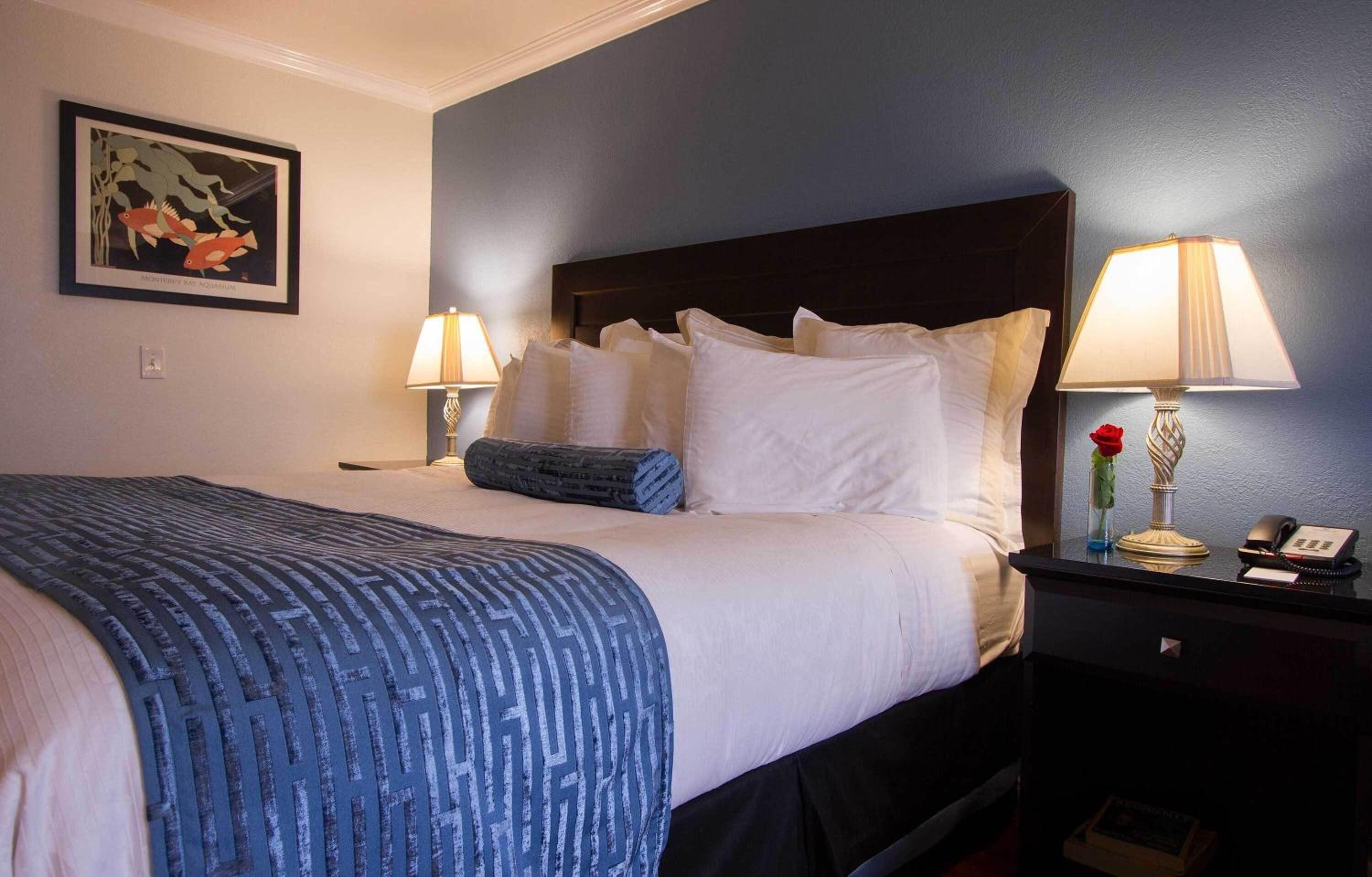 Una cama bien hecha con ropa de cama blanca y azul, dos lámparas de noche, un cuadro en la pared y una mesita con un jarrón y un teléfono.