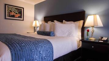 Una cama bien hecha con ropa de cama blanca y azul, dos lámparas de noche, un cuadro en la pared y una mesita con un jarrón y un teléfono.