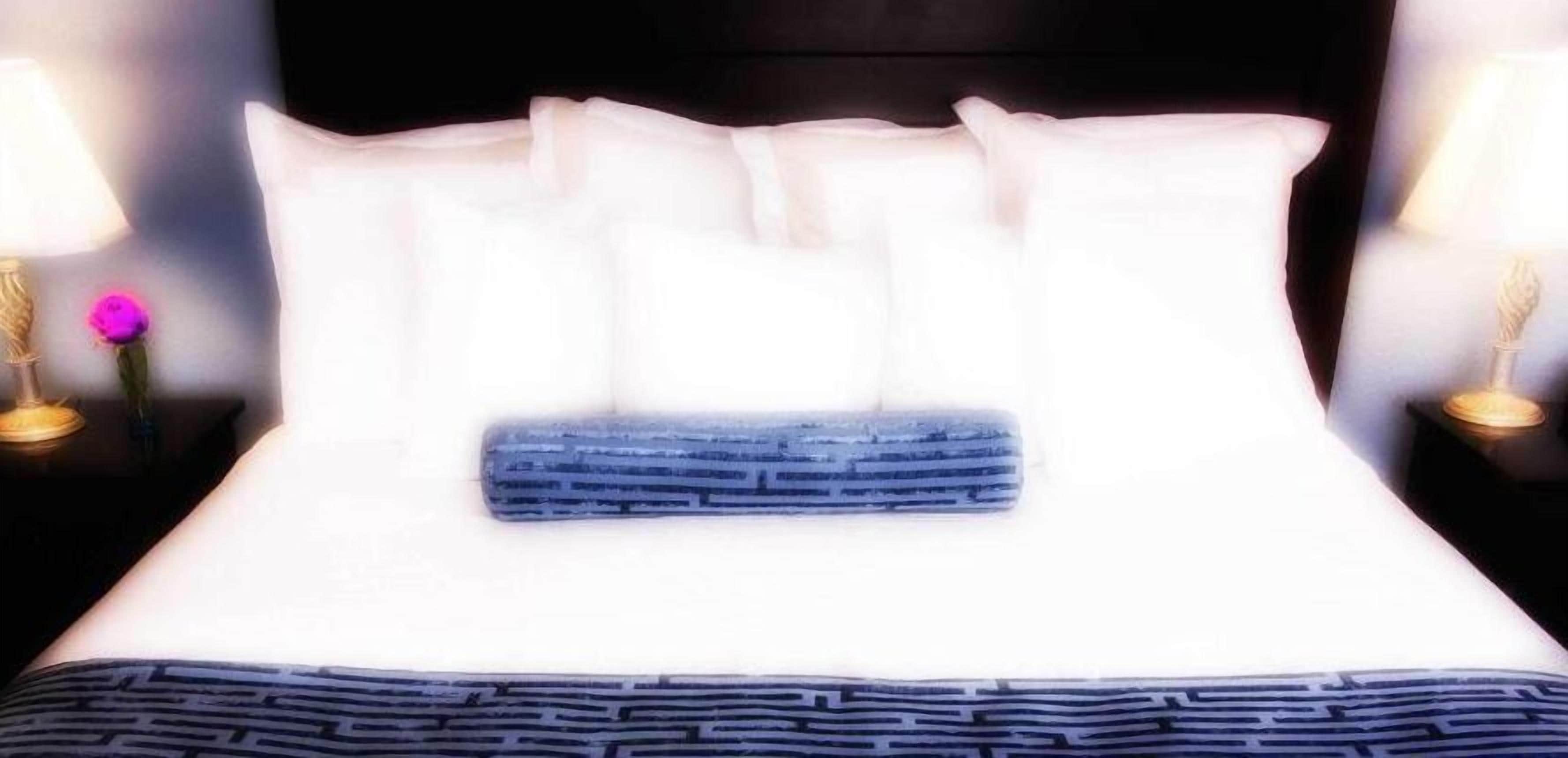 Una cama con almohadas blancas y una manta azul, con dos lámparas de noche adornadas con flores.