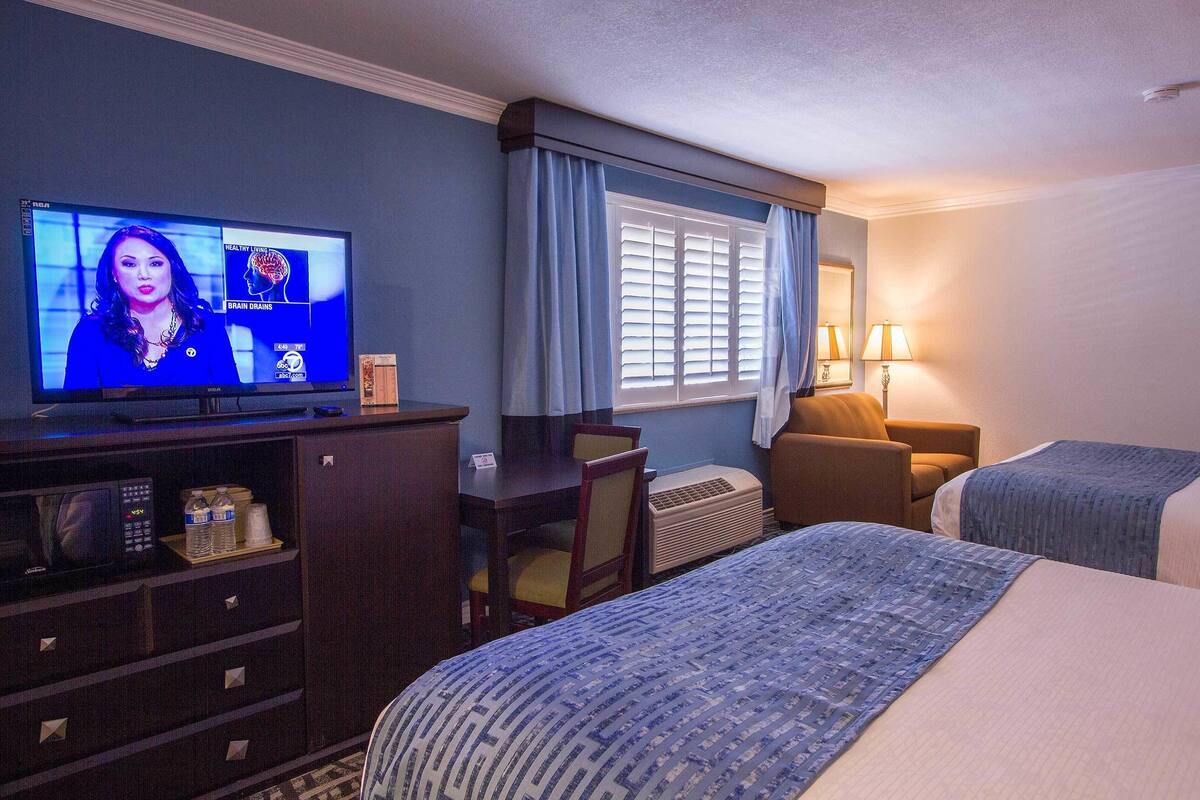 A hotel room with a television displaying a news program, a desk with a chair, a bed with a blue blanket, and a window with white blinds.