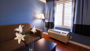 Ein Zimmer mit einem braunen Sofa, einem kleinen Tisch mit einer Vase weißer Blumen, einer Stehlampe und einem Fenster mit Jalousien und Vorhängen.