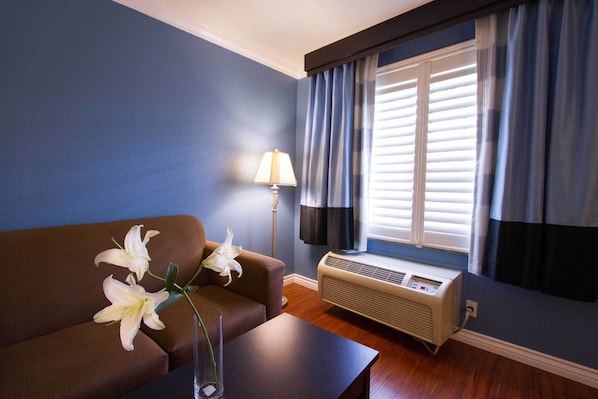 Una habitación con un sofá marrón, una mesita con un jarrón de flores blancas, una lámpara de pie y una ventana con persianas y cortinas.