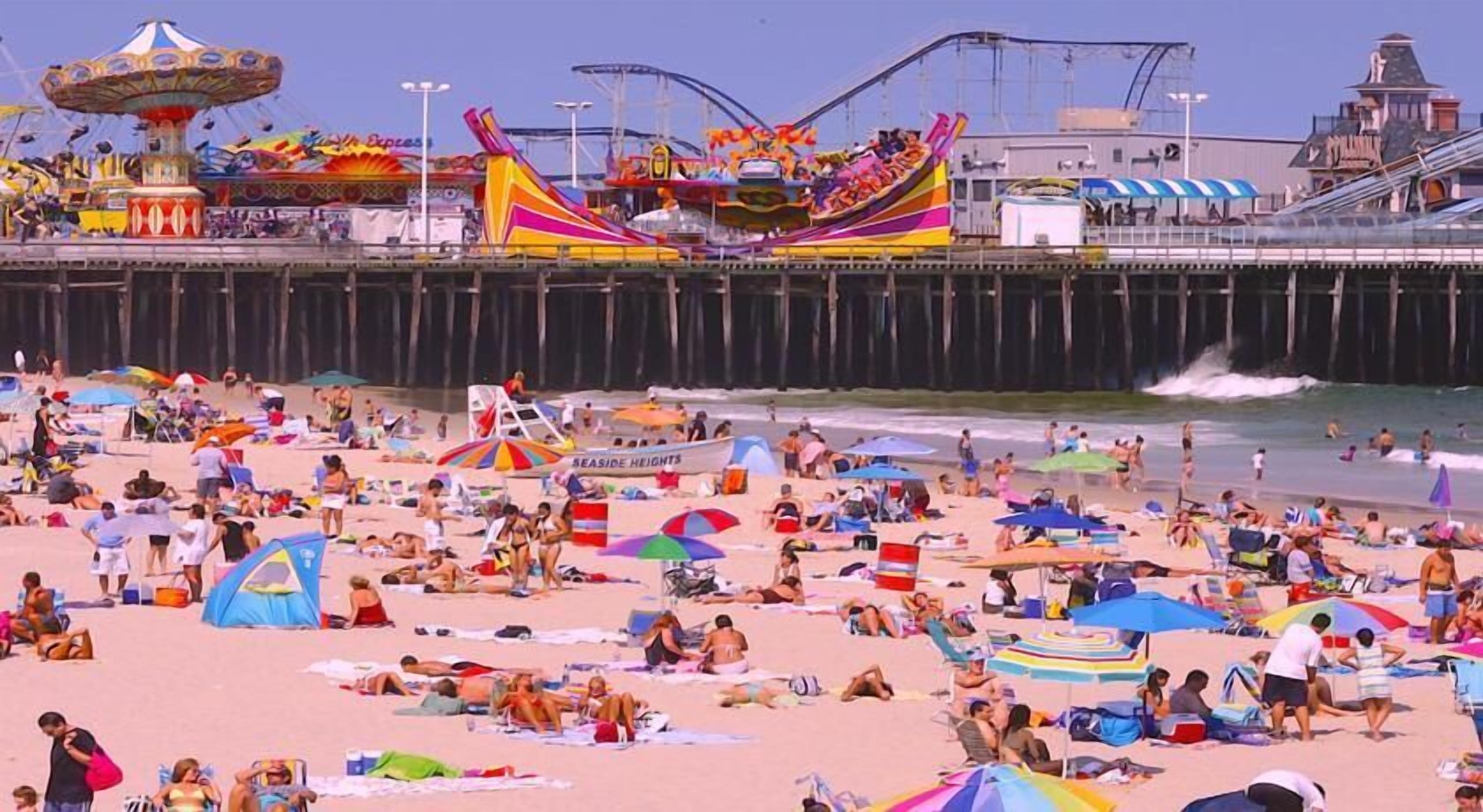 A crowded beach with people sunbathing and swimming, a pier with amusement rides, and a sign for 'SeaSide Heights'.