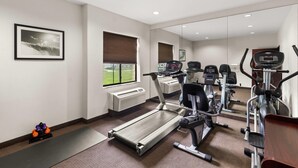 Ein Fitnessraum mit Laufband, Crosstrainer und Gewichten, einem Fenster mit Jalousien und einem Spiegel, der die Geräte reflektiert.