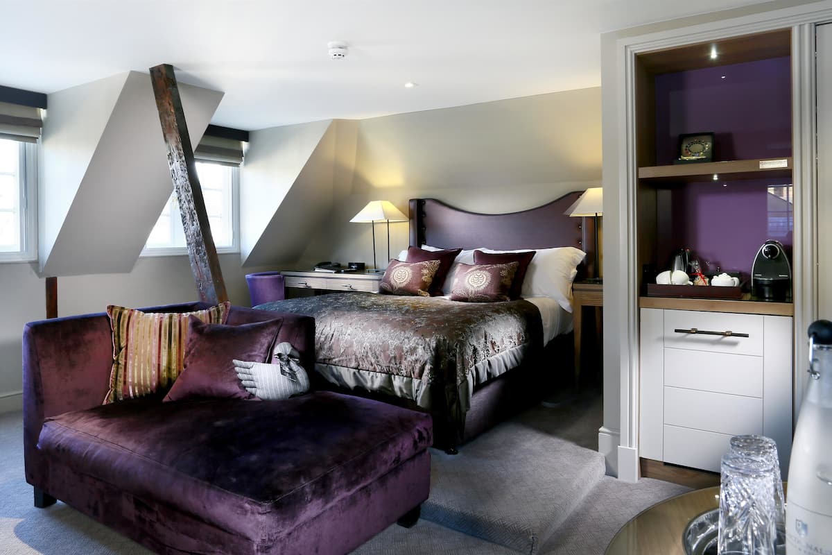 A modern bedroom with a sloped ceiling, a large bed with a headboard, a purple armchair, a bedside table with a lamp, and a wardrobe.