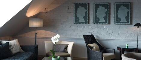 Una sala de estar moderna estilo loft, con techo inclinado, un ventanal grande y pared de ladrillo. Se observan tres retratos enmarcados en la pared, una orquídea en maceta sobre una mesa de vidrio, y diversas opciones de asientos, incluyendo un sofá, sillones y una butaca.