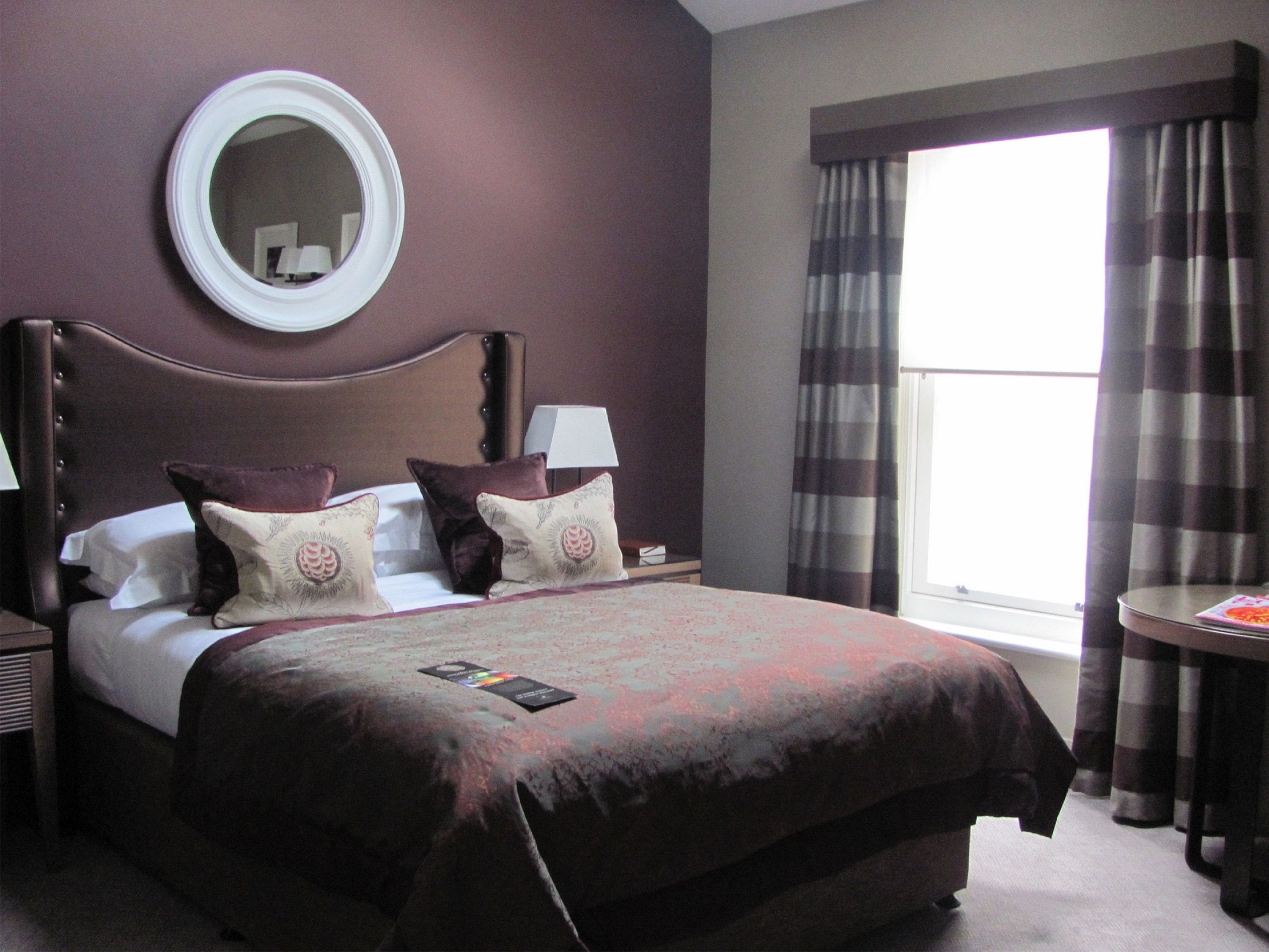 A hotel room with a large bed, two bedside tables, a round mirror, a window with curtains, and a small round table.
