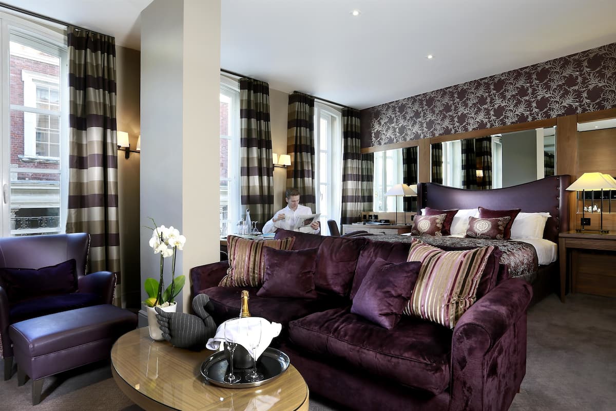 A hotel room with a large bed, a purple sofa, a coffee table, and a view of a cityscape through the windows.