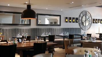 A restaurant with a modern interior, featuring a brick wall, a large mirror, and a round clock. Tables are set with glasses and cutlery.