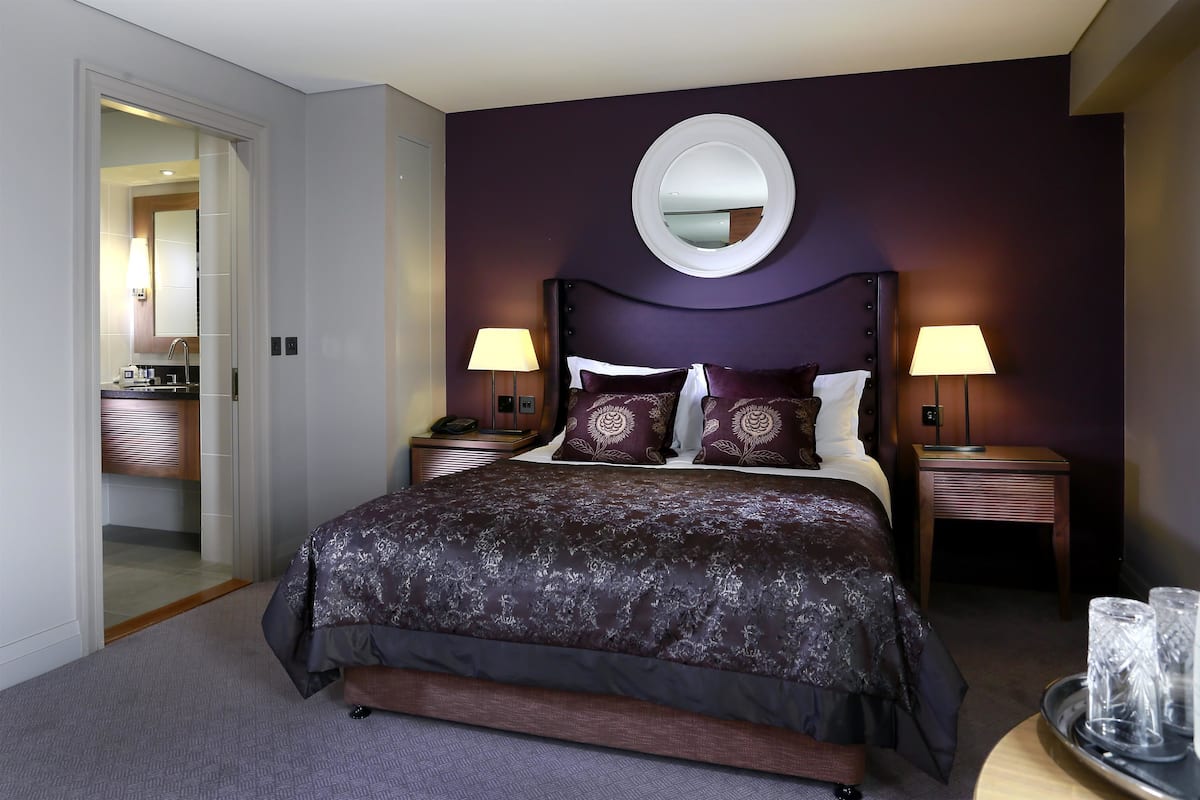 A hotel room with a large bed, two bedside tables with lamps, a round mirror on the wall, and a small table with glasses and a bottle.