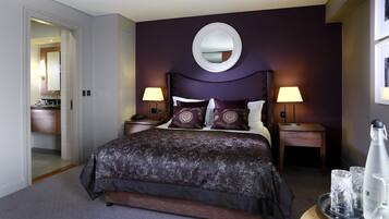 A hotel room with a large bed, two bedside tables with lamps, a round mirror on the wall, and a small table with glasses and a bottle.