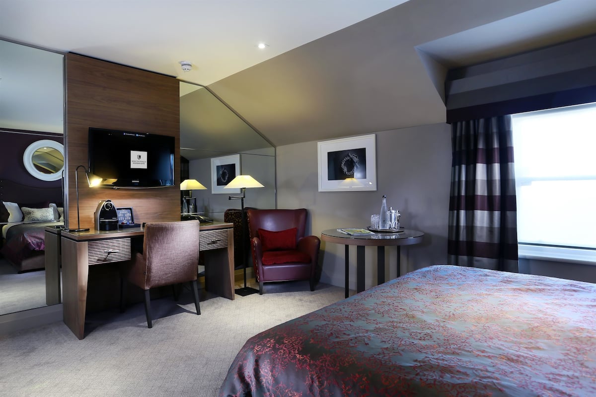 A hotel room with a bed, desk, chair, TV, and a small table with a lamp.