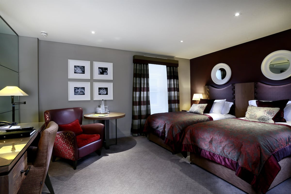 A hotel room with two beds, a desk, a chair, a lamp, and a mirror.