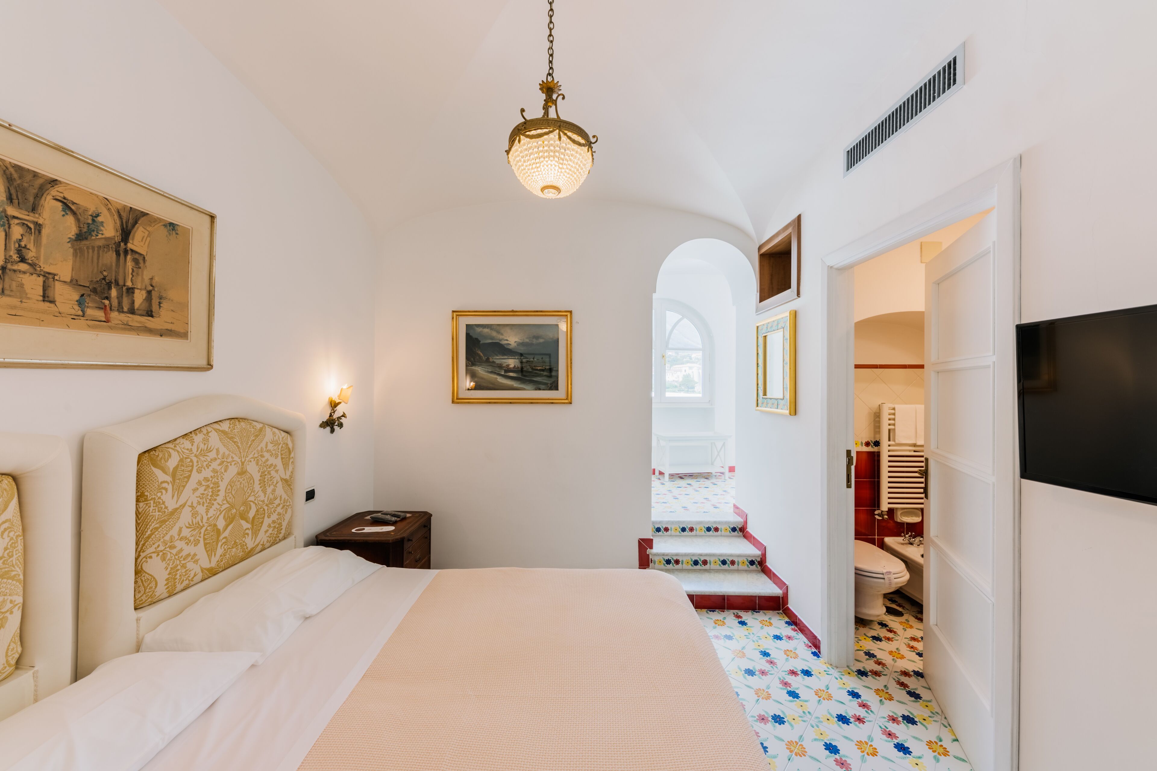 A bedroom with a bed, a nightstand, a wall-mounted lamp, a painting, and a view of the bathroom through an open door.