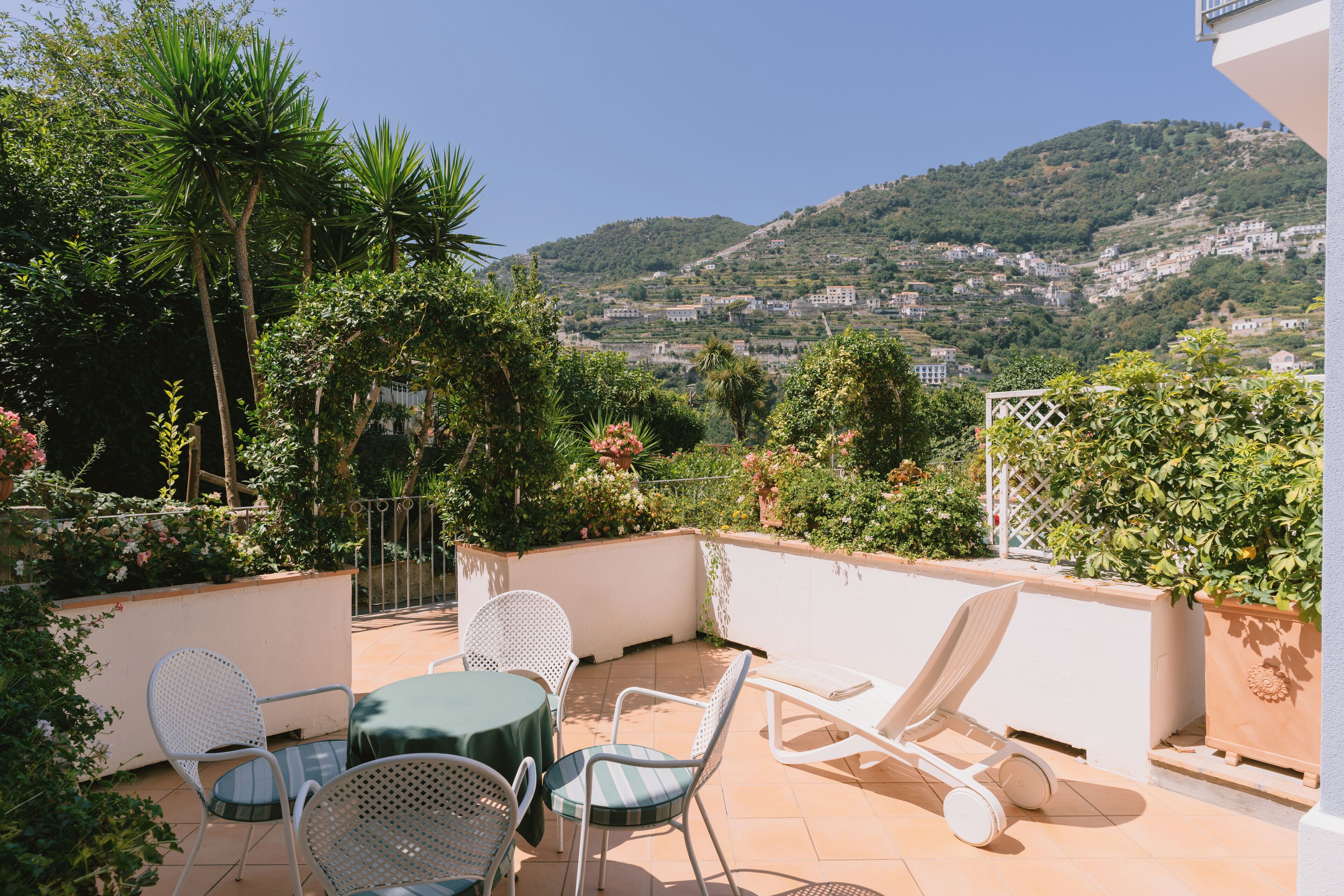 A terrace with a table and chairs, surrounded by plants and overlooking a hillside with buildings.