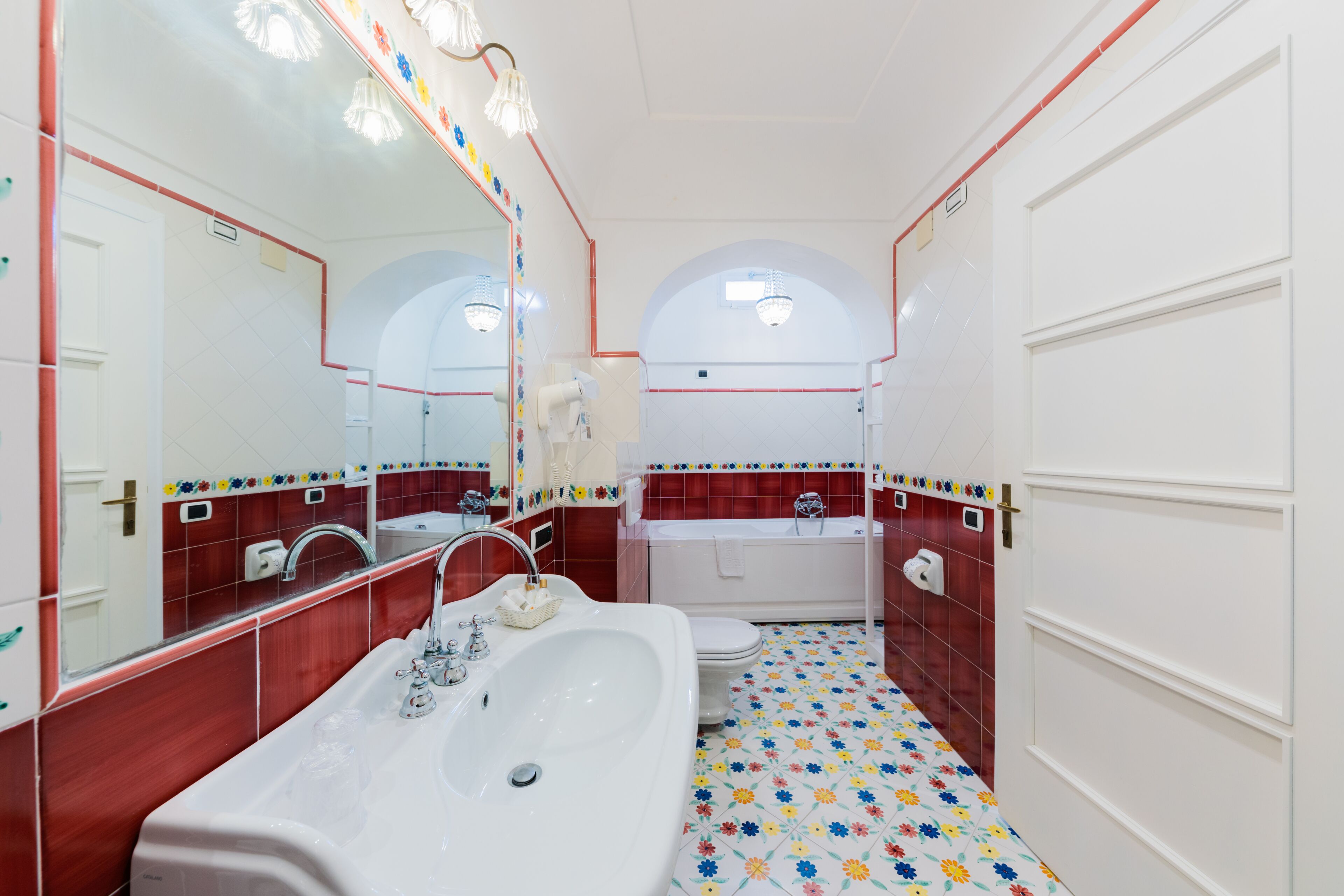 A bathroom with a large white sink, a mirror, and a bathtub. The floor has a colorful floral pattern.