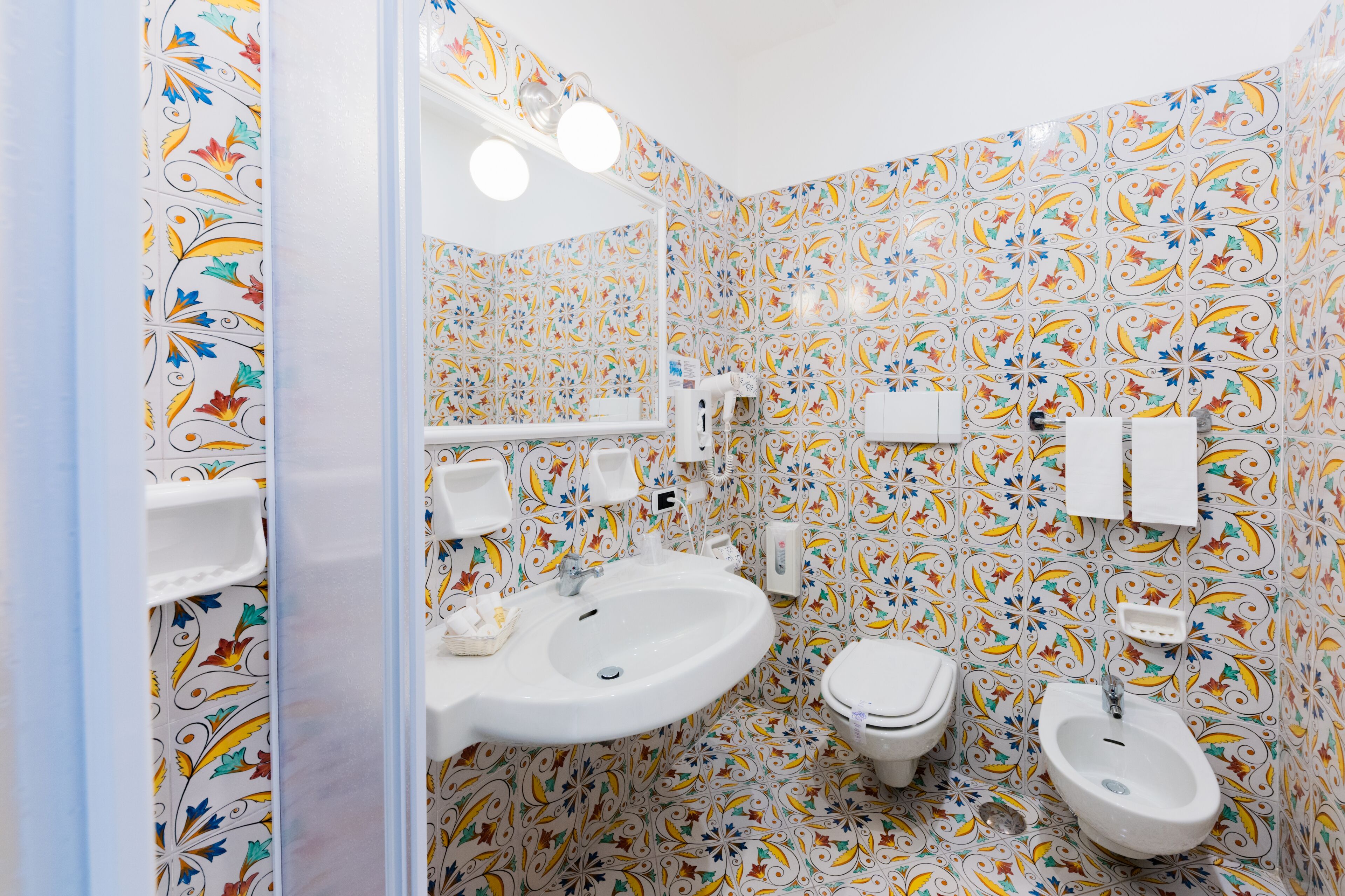 Un baño con lavamanos, inodoro y bidé, todo con sanitarios blancos. Las paredes están cubiertas de azulejos florales de colores.