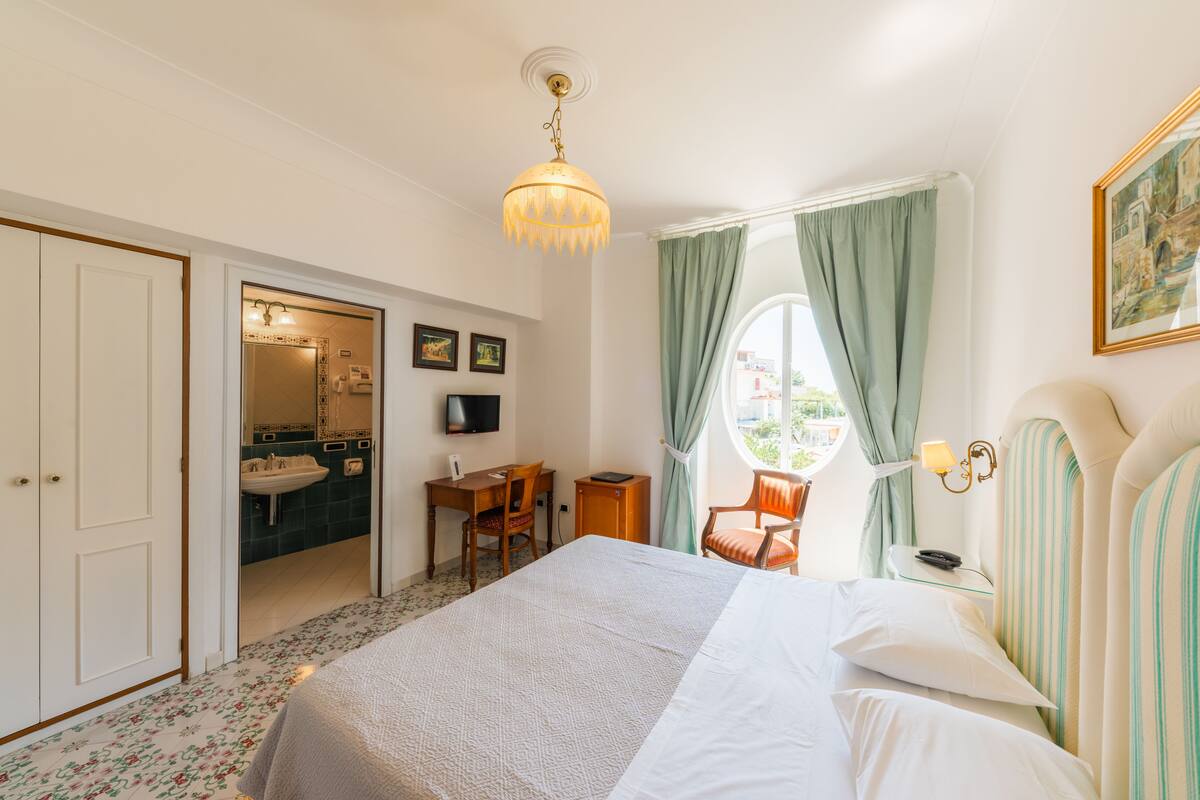 A hotel room with a bed, a desk, a chair, a window with curtains, a painting on the wall, and a bathroom visible through an open door.
