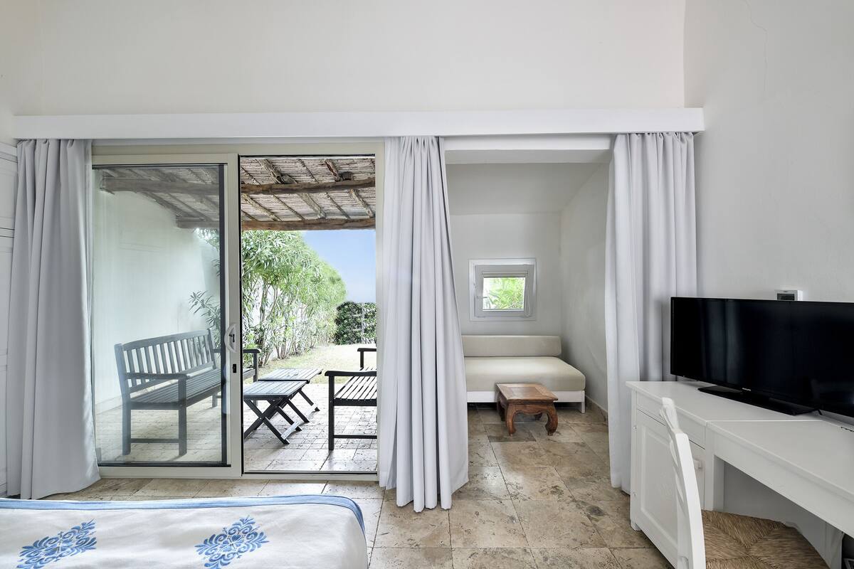A hotel room with a bed, a TV, a sofa, a small table, and a sliding glass door leading to an outdoor patio with a bench and a table.