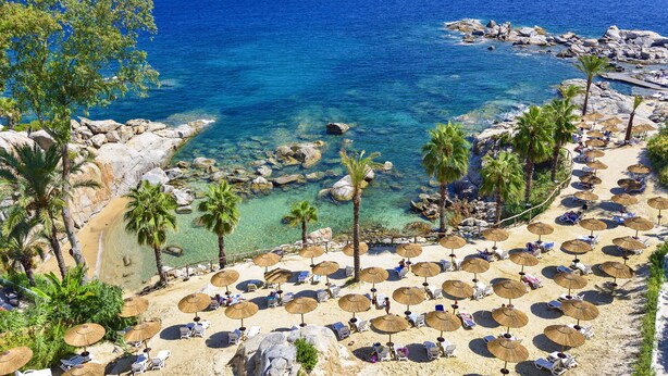 A beach with clear blue water, rocky shoreline, palm trees, and sun loungers with umbrellas.