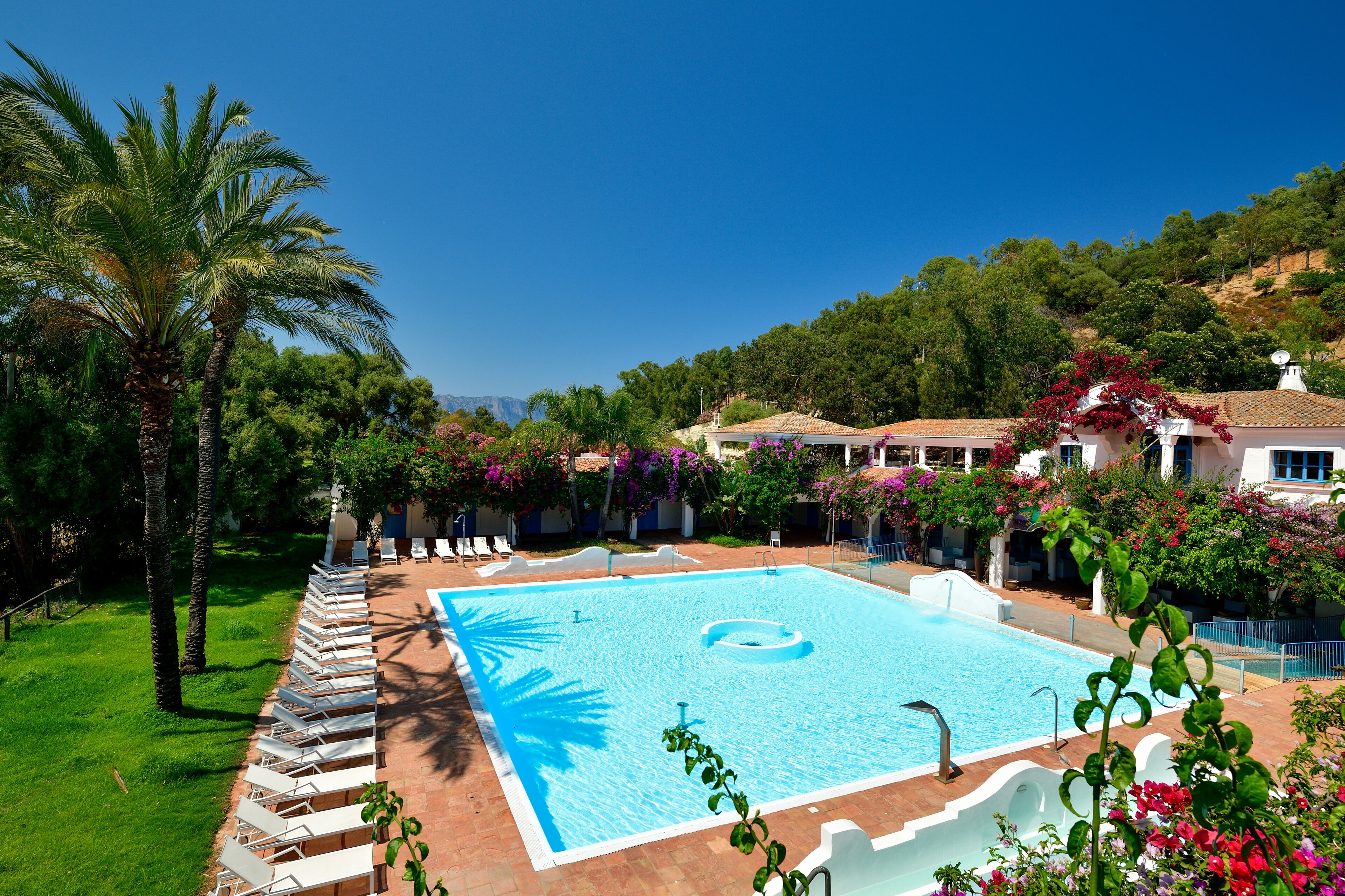 Ein Pool mit Liegestühlen und Palmen, umgeben von Grünflächen und Gebäuden.