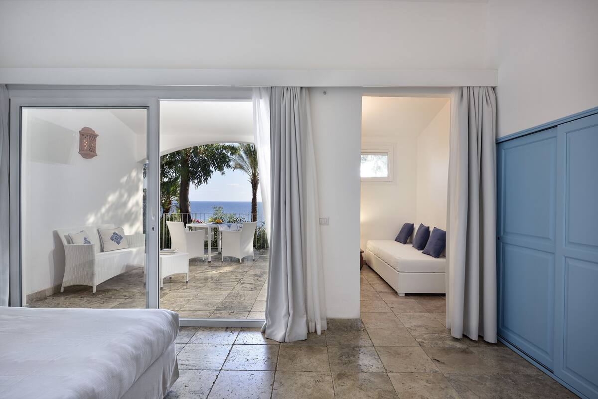 A hotel room with a bed, a seating area with a sofa and chairs, and a view of the sea through a glass door.