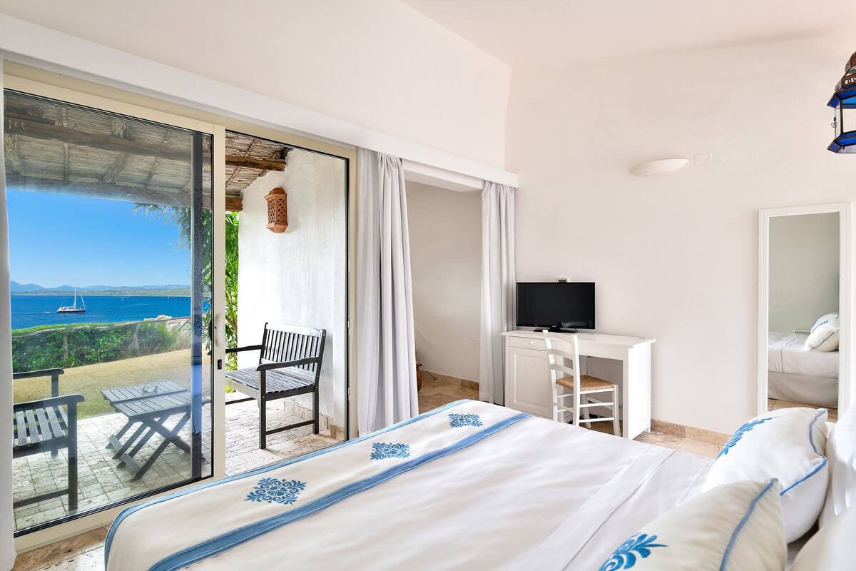 A hotel room with a bed, a desk, a TV, and a balcony with a view of the sea.