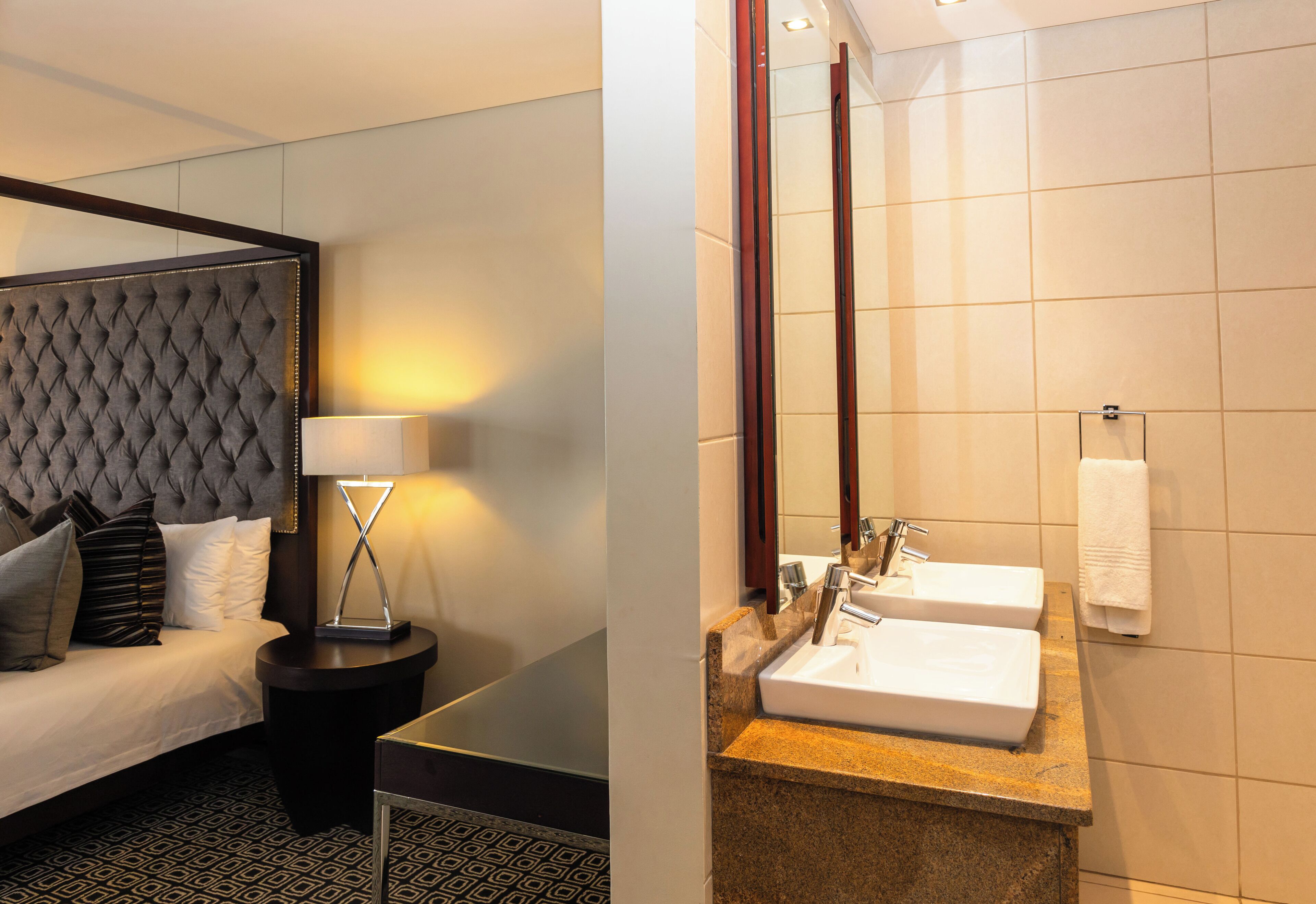 A hotel room with a tufted headboard, a bedside table with a lamp, a bed with pillows, and a bathroom with two sinks and a mirror.
