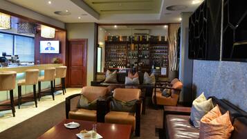 Un rincón de bar con una pantalla de televisión, asientos con cojines y una estantería para vinos al fondo.