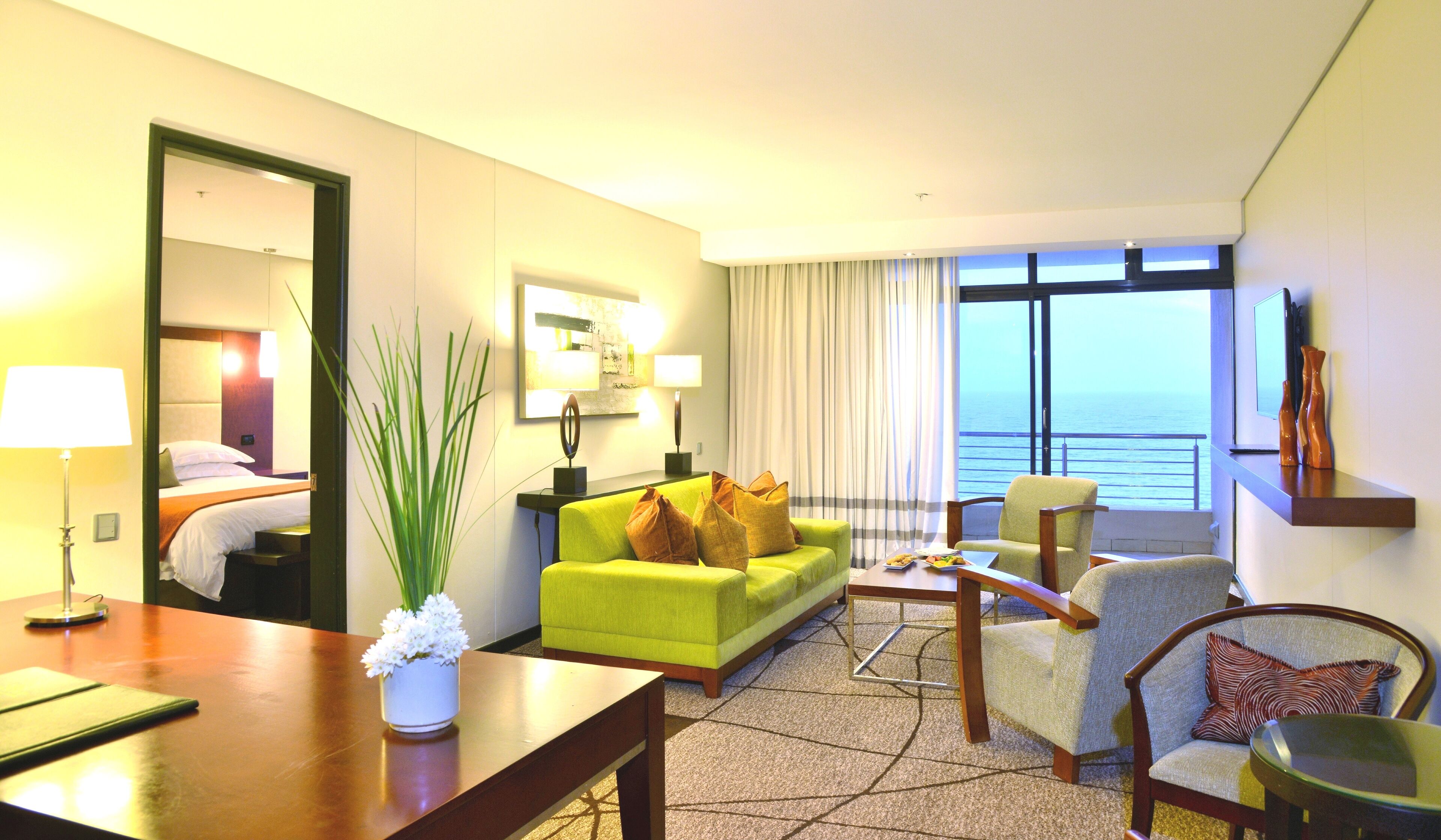 A hotel room with a green sofa, a wooden coffee table, a chair, a desk, and a view of the ocean.