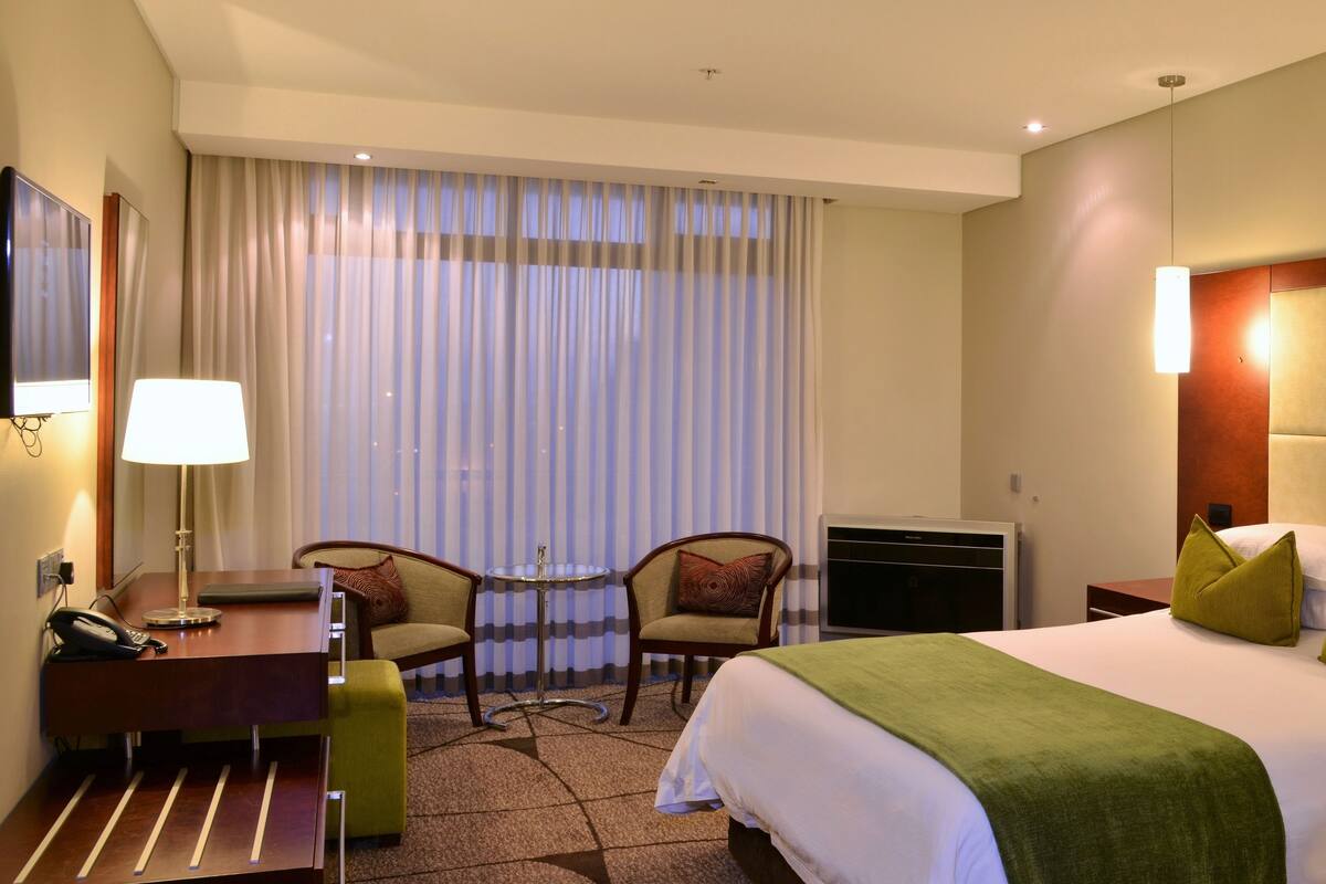 Une chambre d’hôtel avec un lit, un bureau avec une lampe, deux chaises, un téléviseur et une grande fenêtre avec des rideaux.