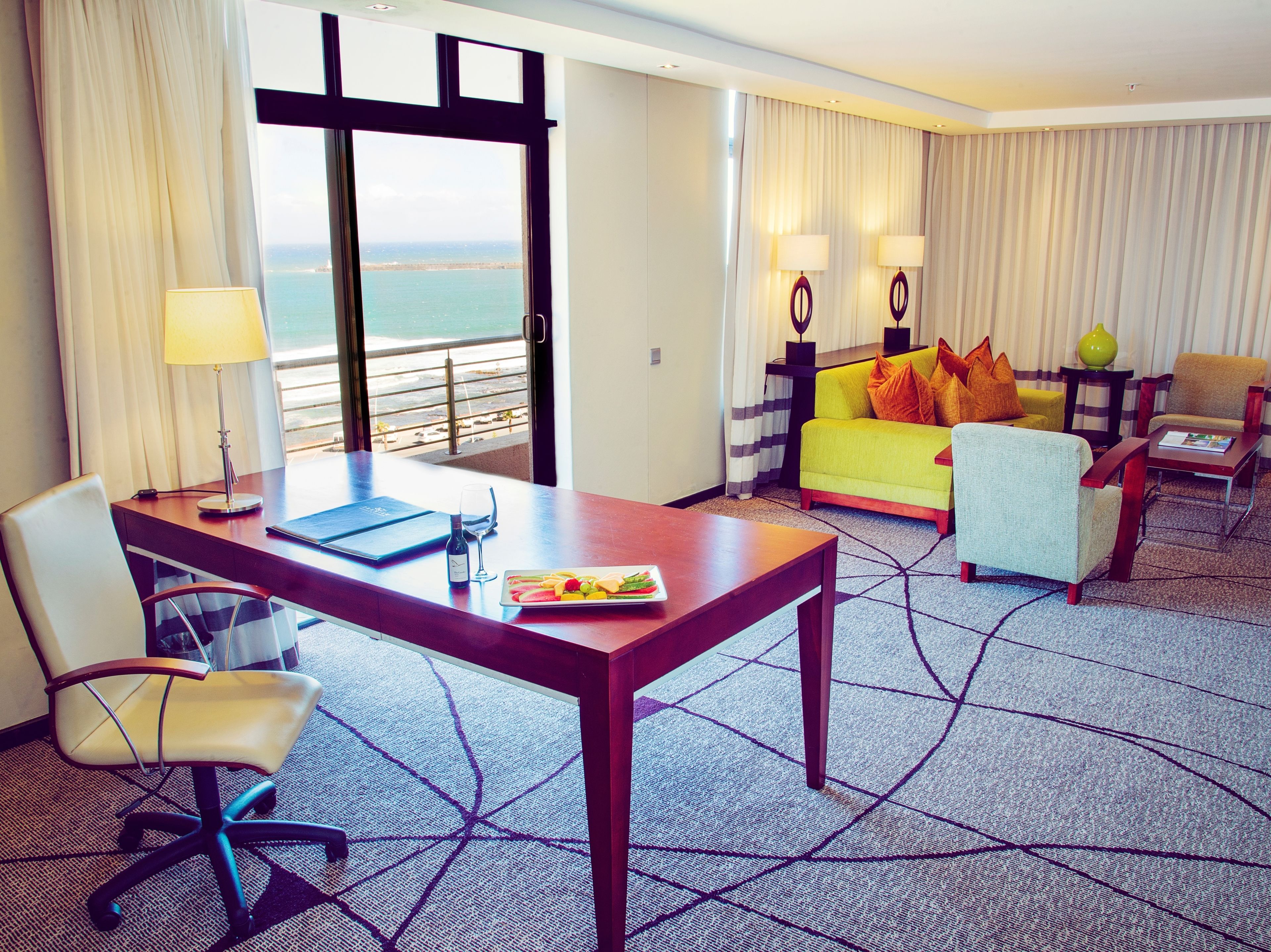 Habitación de hotel con una mesa de comedor, un sofá, una silla, una lámpara y vista al océano.