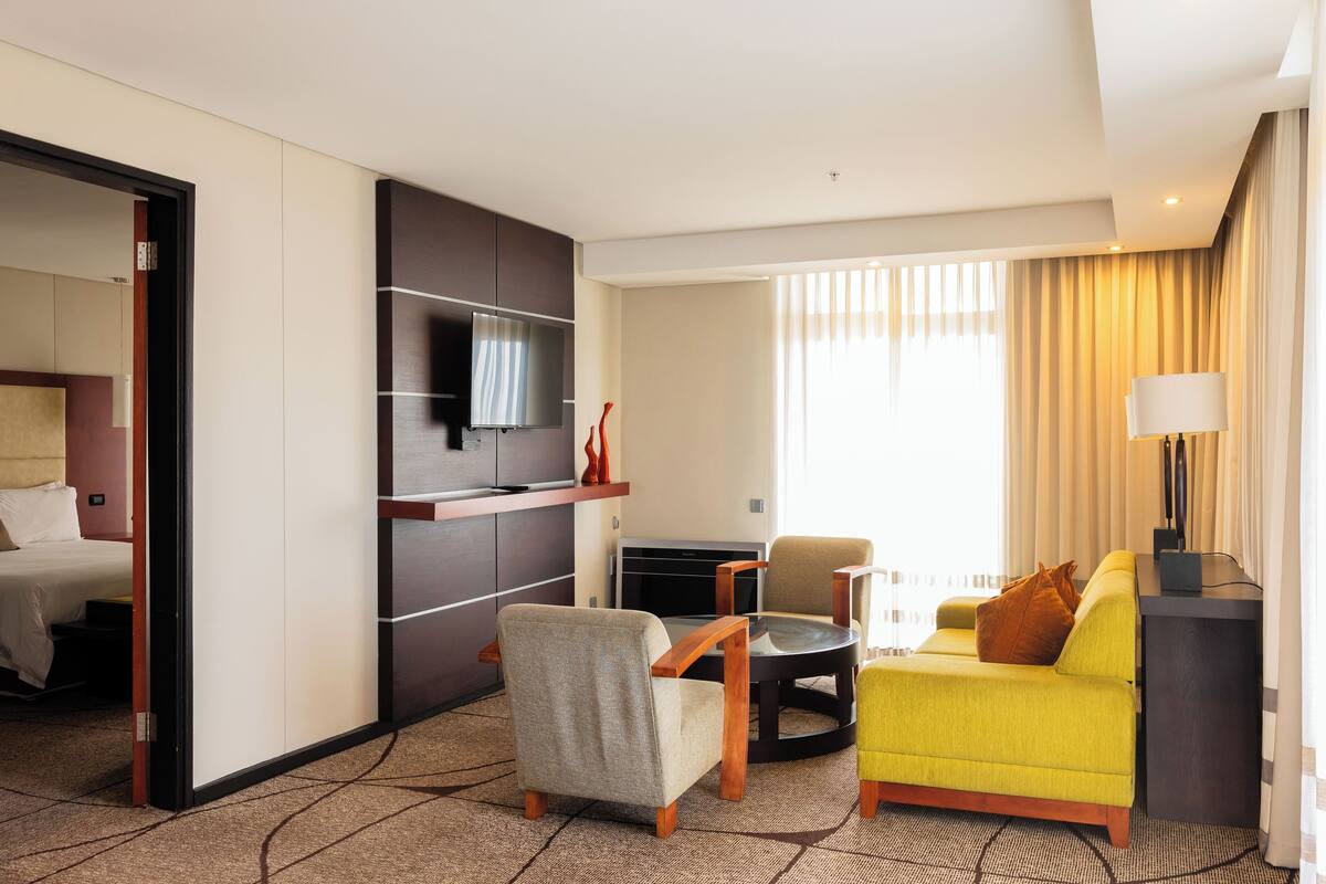 Une chambre d’hôtel avec un lit, un canapé jaune, un fauteuil gris, une petite table ronde, une télévision et un bureau avec une lampe.