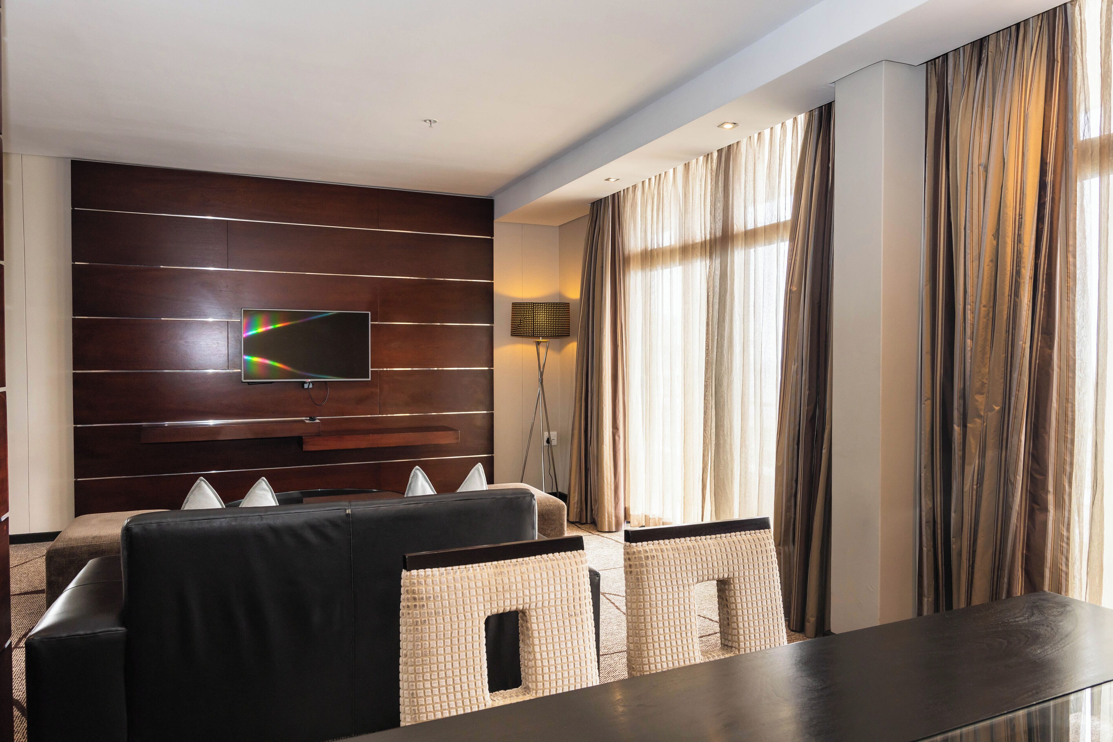Une chambre d’hôtel moderne dotée d’un téléviseur à écran plat fixé sur un panneau mural en bois, d’un canapé, d’une table à manger et d’une lampe.