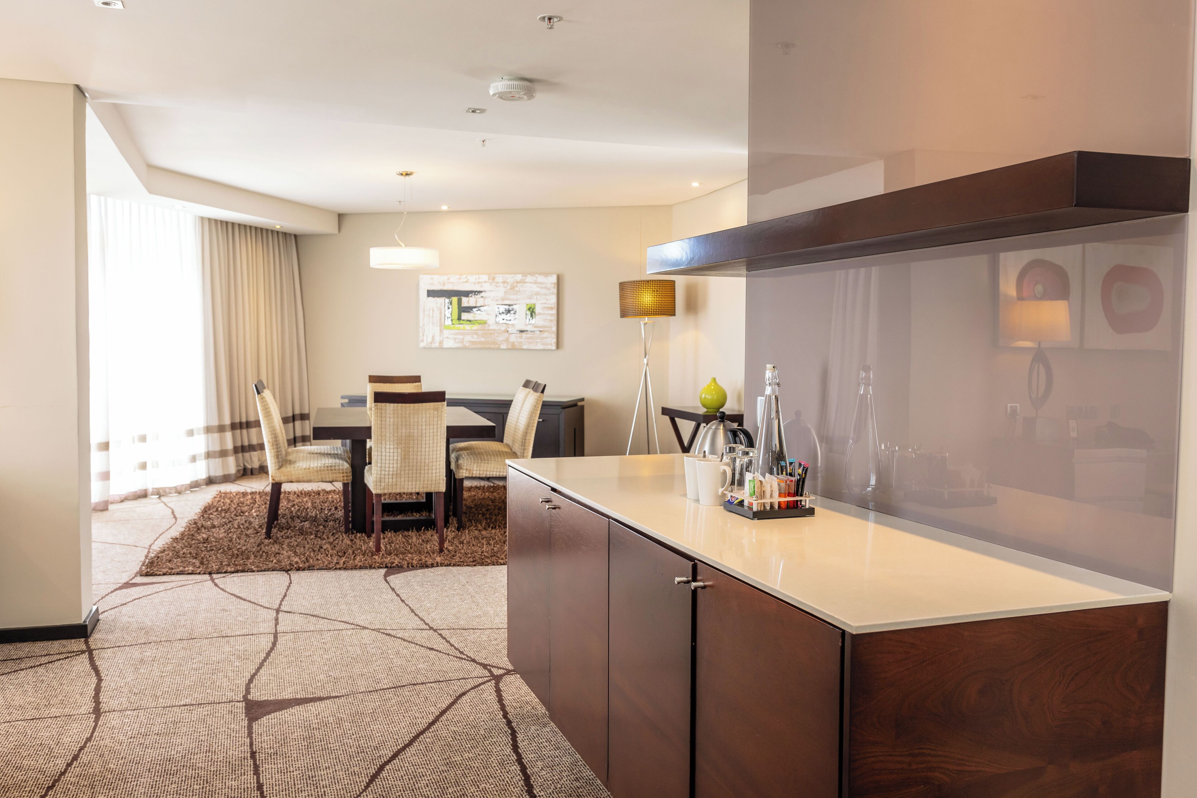 A modern hotel room with a dining area, a large window with curtains, a flat-screen TV, and a minibar.