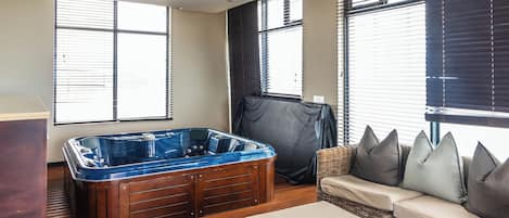 Une pièce dotée d'un plancher en bois, d'un grand jacuzzi, d'un canapé en osier avec des coussins et de grandes fenêtres équipées de stores.