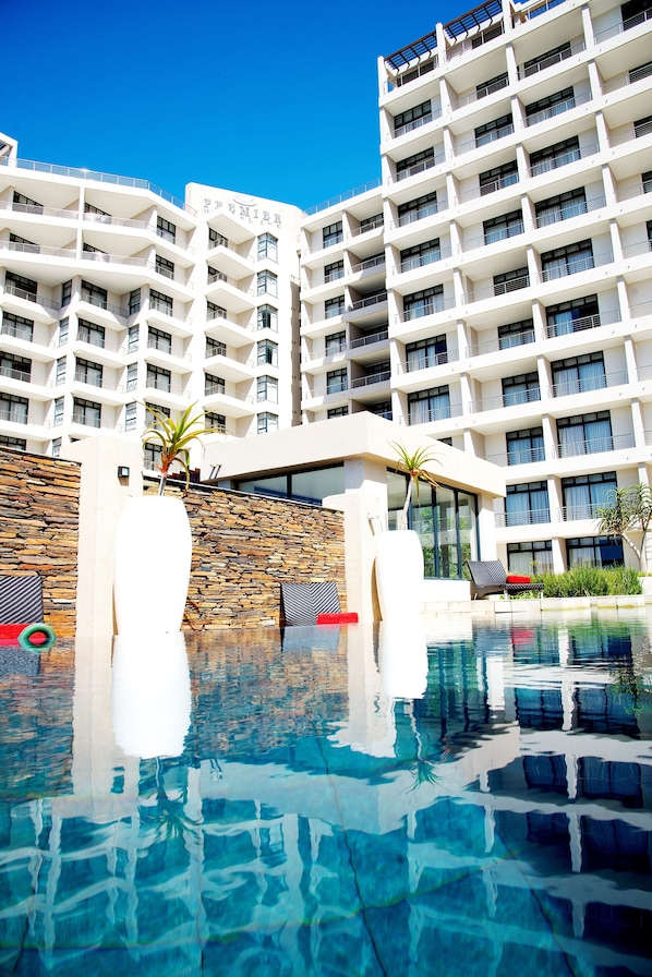 A modern hotel with a pool area, featuring a stone wall, lounge chairs, and a clear blue sky.