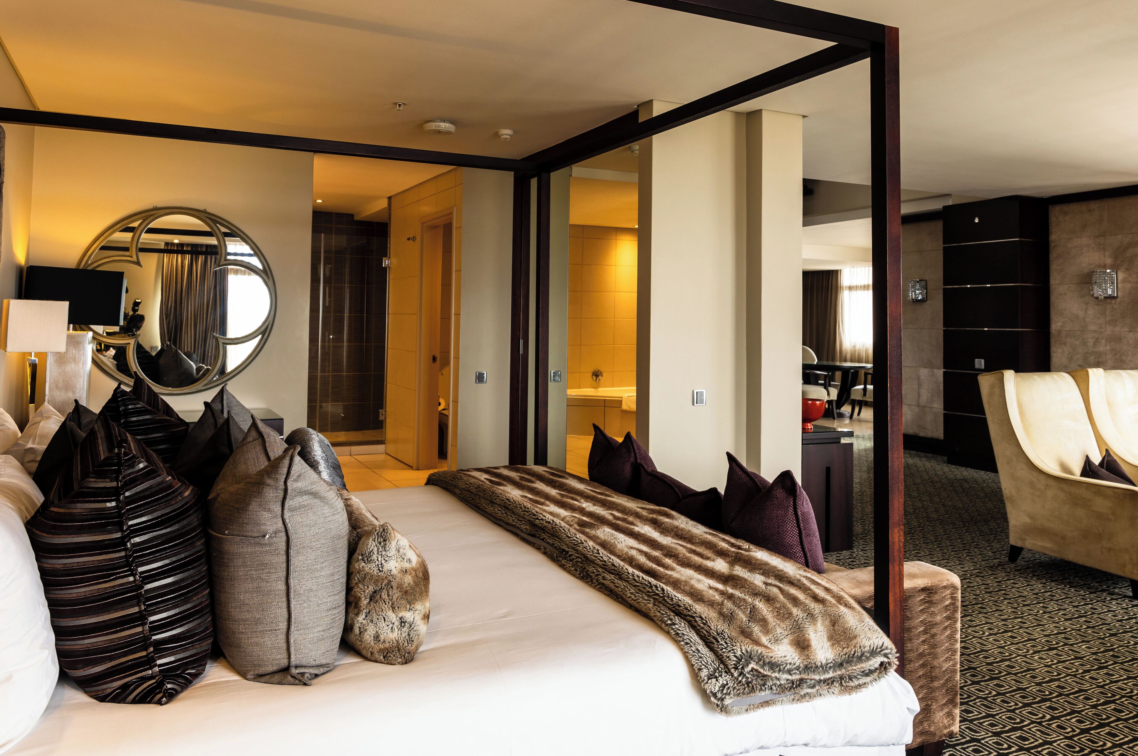 A hotel room with a large bed, a sitting area with a sofa, a dining table, and a mirror.