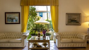 A room with two white upholstered sofas, a glass-top coffee table with decorative items, yellow curtains, and framed artwork on the walls. - Hotel Sylvia (Camaiore)