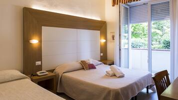 A hotel room with two beds, a wooden headboard, bedside lamps, a small table, and a view of greenery through the window.