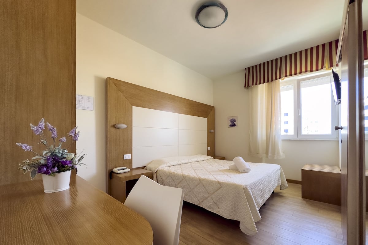 A hotel room with a wooden headboard, two beds, a wooden table, a chair, a potted plant, a bedside table with a phone, and a window with curtains.