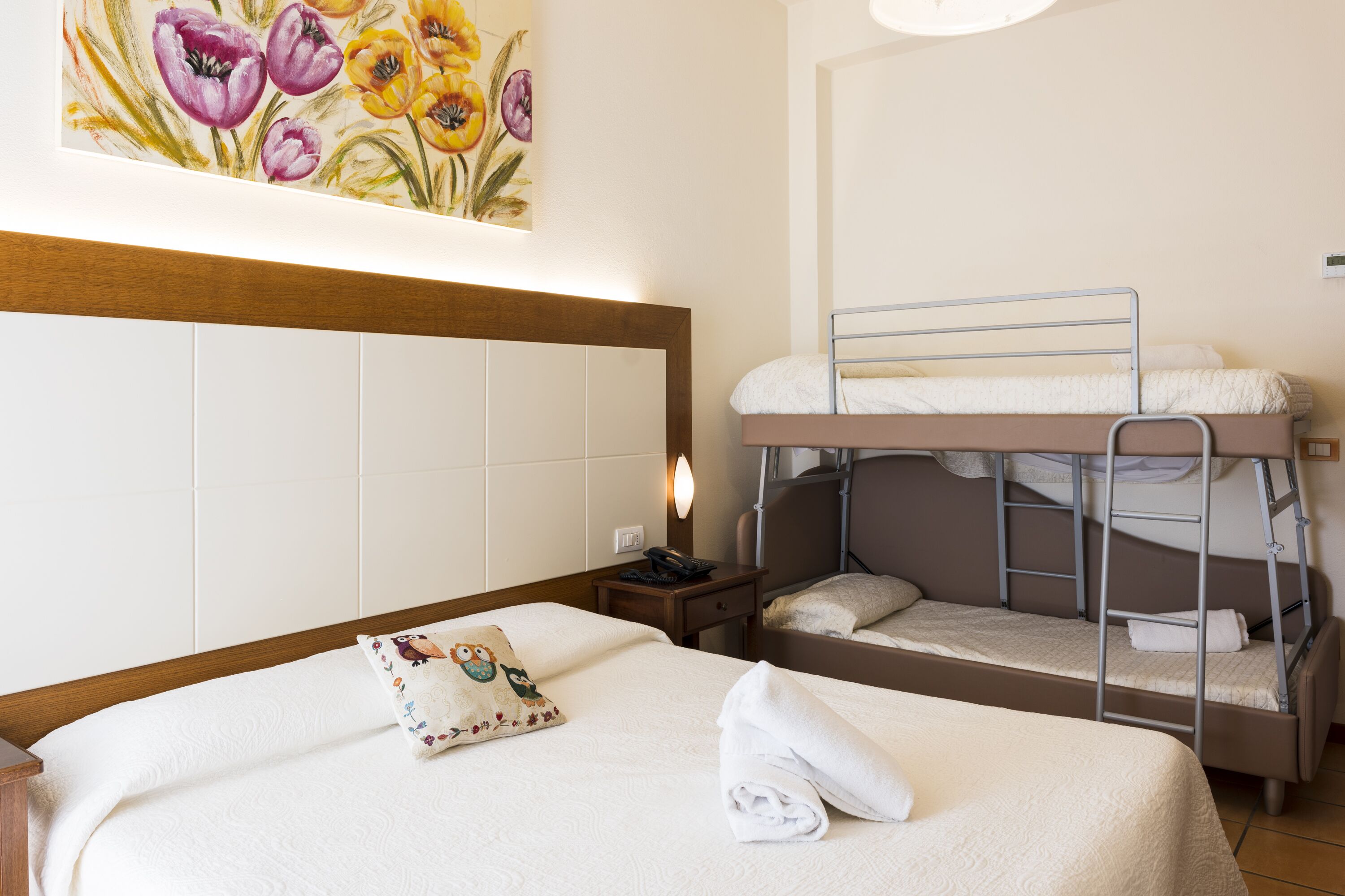 Une chambre d’hôtel avec un lit simple, un lit superposé, une table de chevet avec une lampe et un tableau représentant des fleurs accroché au mur.