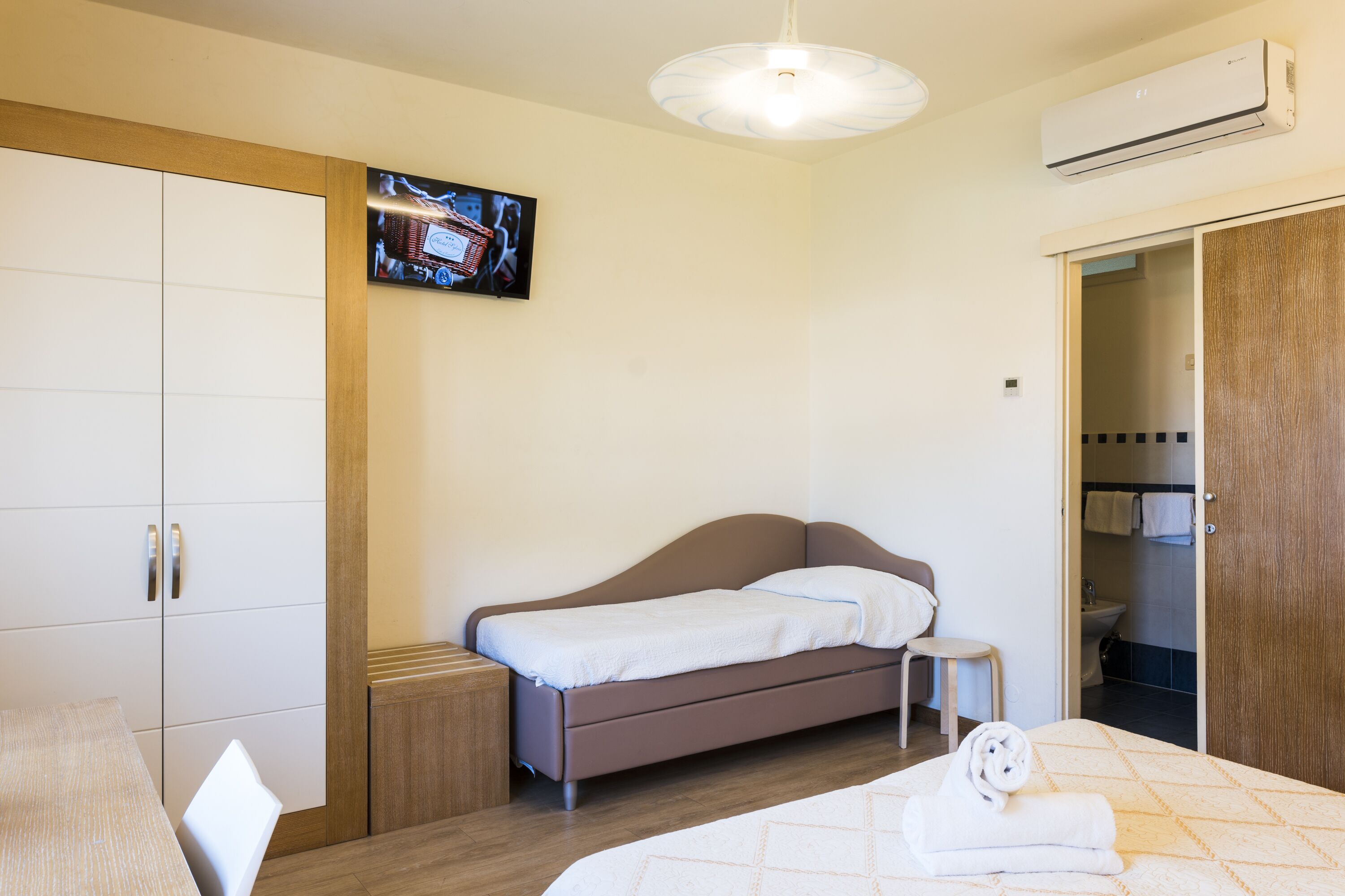 A bedroom with a single bed, a small table, a TV mounted on the wall, and a wardrobe.