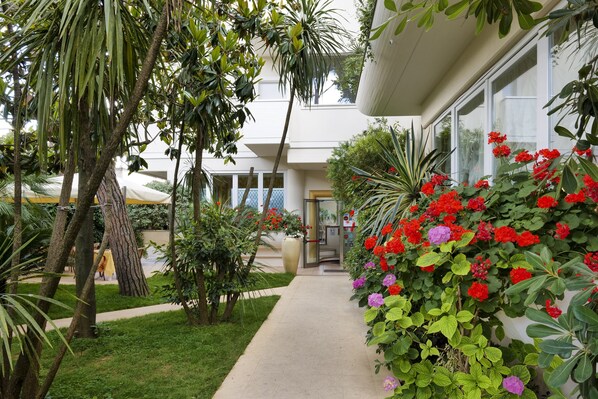 A pathway leading to a building with a glass door, surrounded by lush greenery and flowering plants.